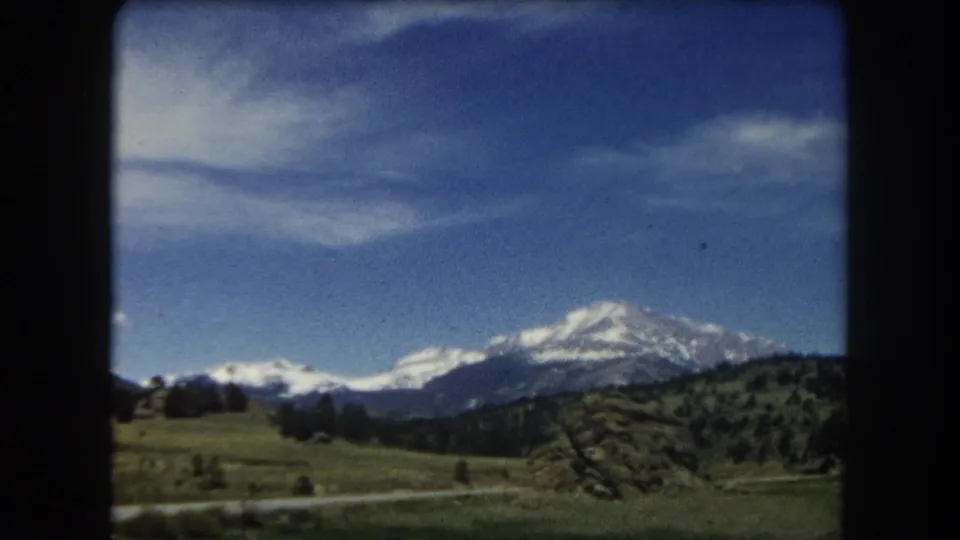 End-of-pan 8mm frame from Summit County, Colorado, 1962; snow-covered mountain range over open green hills and scattered conifers under blue sky.