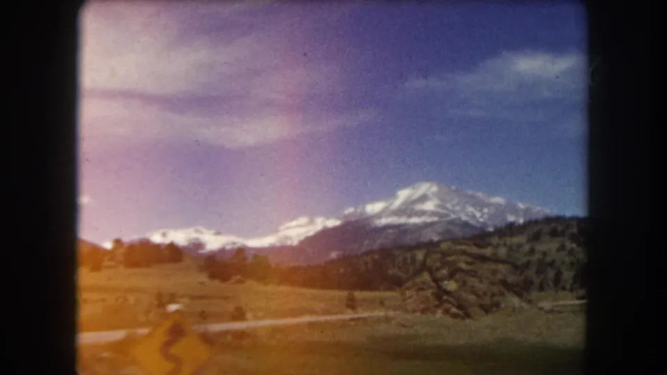 Wide 8mm landscape shot of a snow-capped mountain peak over a meadow and winding road, Summit County, Colorado, circa 1962.