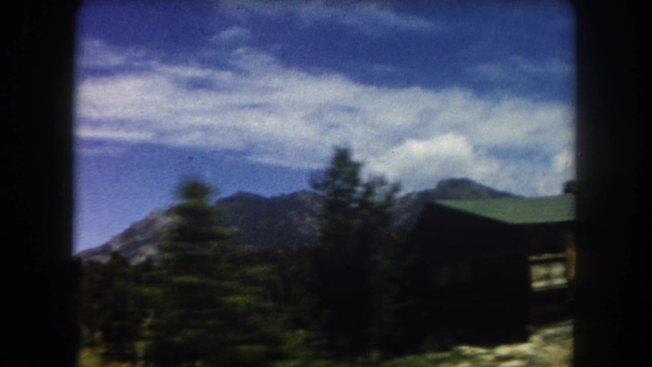 Mountain ridge and pine trees with the corner of a dark-roofed structure visible at right, filmed on 8mm color stock.