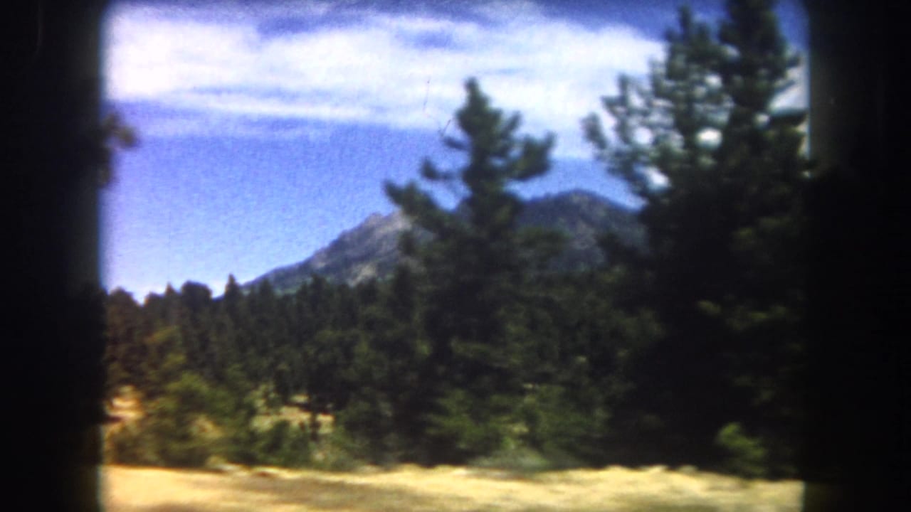 View through car window of pine trees and a distant rocky mountain peak under blue sky, 8mm film grain visible.