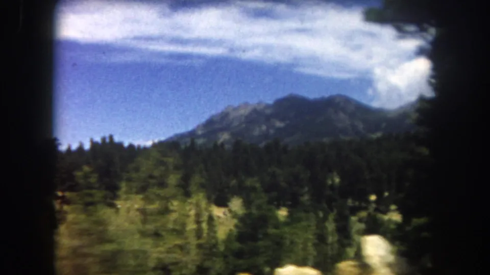 8mm home-movie frame, 1962, Summit County, Colorado: rocky mountain peak, dense subalpine forest, and building cumulus clouds.