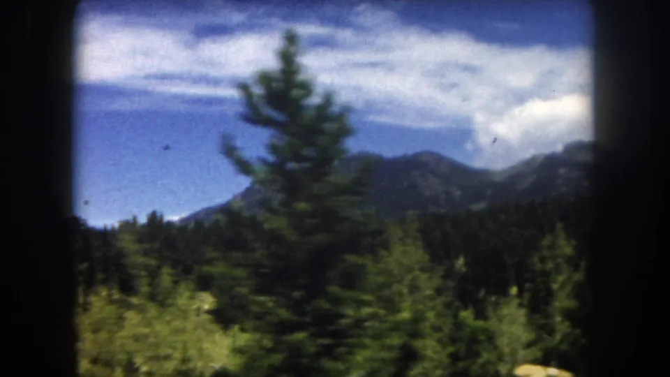 8mm home-movie frame, 1962, Summit County, Colorado: mountain ridgeline, scattered clouds, and conifer trees in the foreground.