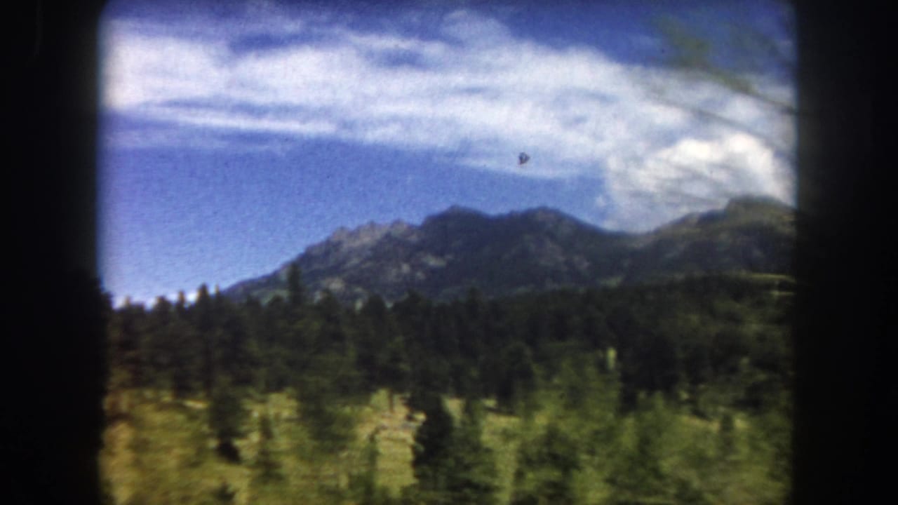 Rocky mountain peaks with dense evergreen forest under a blue sky with scattered clouds, viewed from a moving vehicle; 8mm film with dark frame borders.