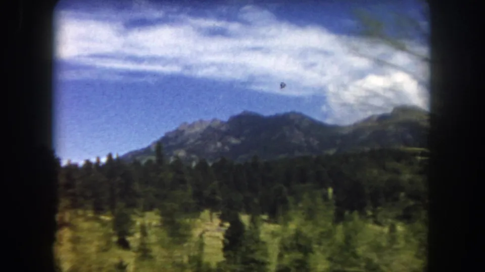 Rugged Colorado Rocky Mountain peaks above a dense evergreen forest, shot from a moving vehicle in 8mm color film, circa 1962.