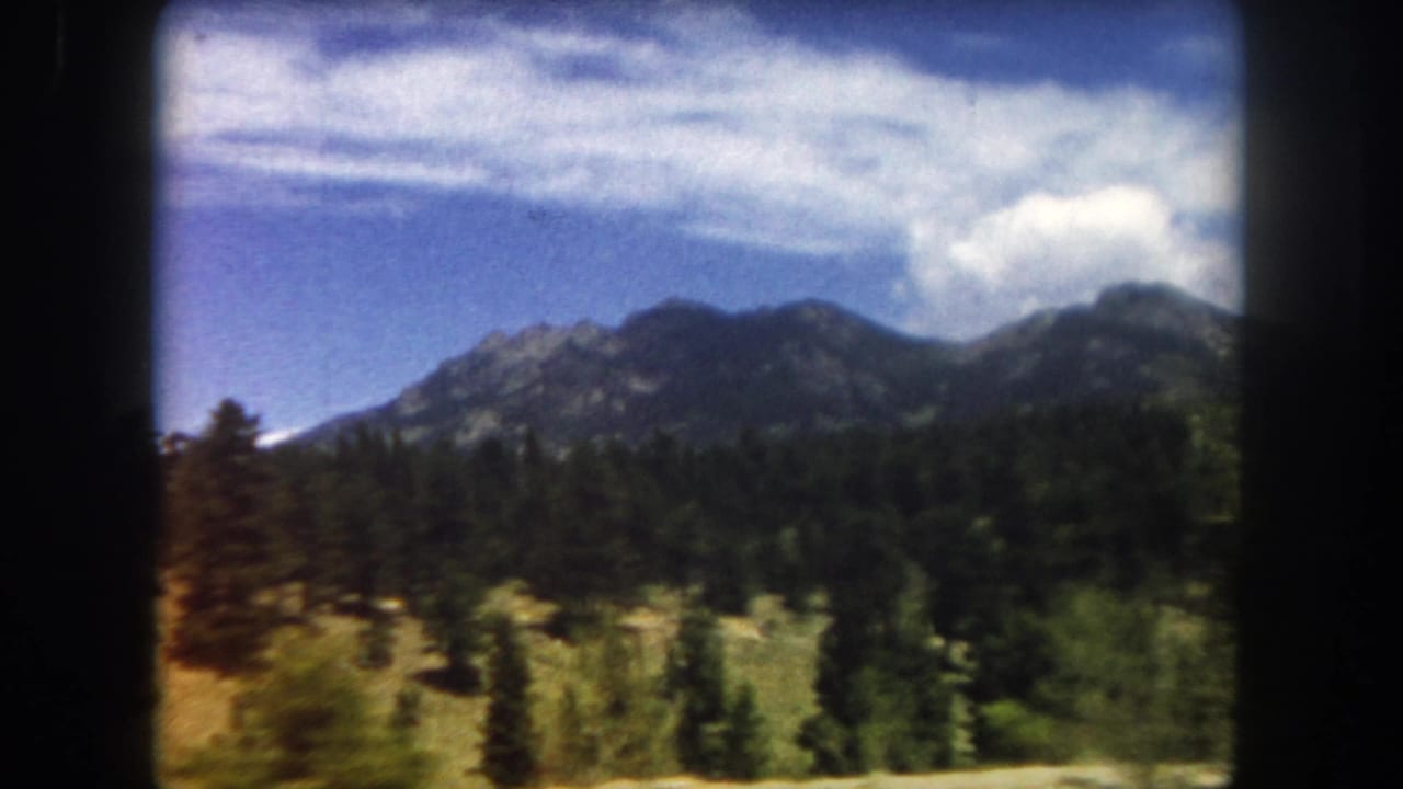 Mountain peaks and dense evergreen forest from a moving vehicle, with blue sky and clouds; warm-toned foliage visible at left; 8mm dark borders.