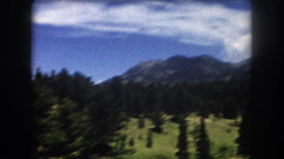 A rounded mountain peak above open meadow and evergreen treeline, filmed from a moving vehicle in Summit County, Colorado, circa 1962.