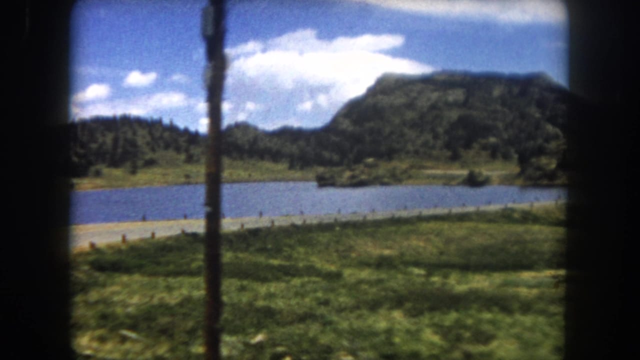 8mm home movie through a car window: calm lake, roadside wooden fence posts, green meadow, and flat-topped mesa, Summit County Colorado, circa 1962.