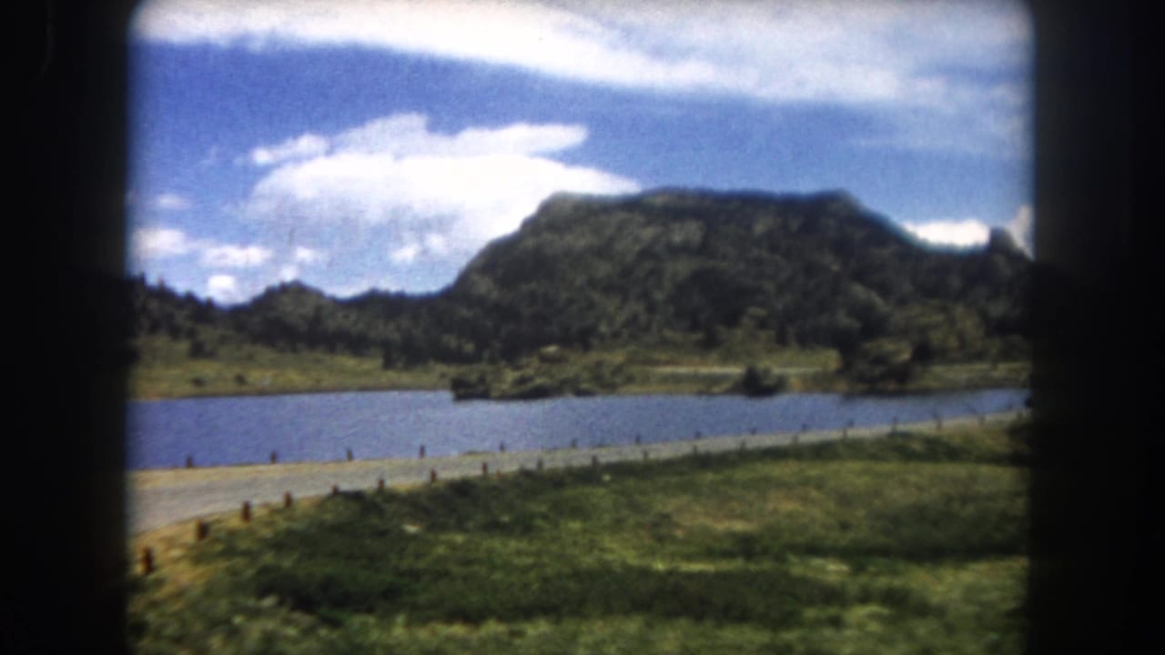Mountain lake, road with fence posts, and flat-topped mesa under a cloudy blue sky, framed by a car window, 8mm film.