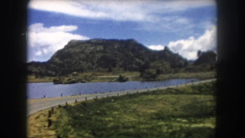 8mm road-trip shot through a car window: alpine lake, fence-post road edge, meadow, flat mesa, and a rocky outcrop to the right, Summit County Colorado, circa 1962.
