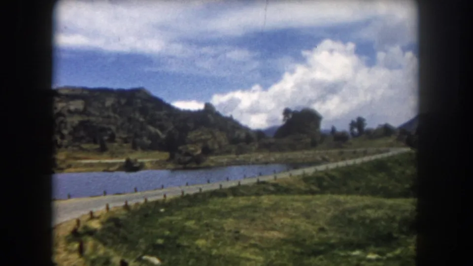 Closing 8mm frame through a car window: lake, road, green meadow, flat mesa, and a rocky peak emerging at right, Summit County Colorado, circa 1962.