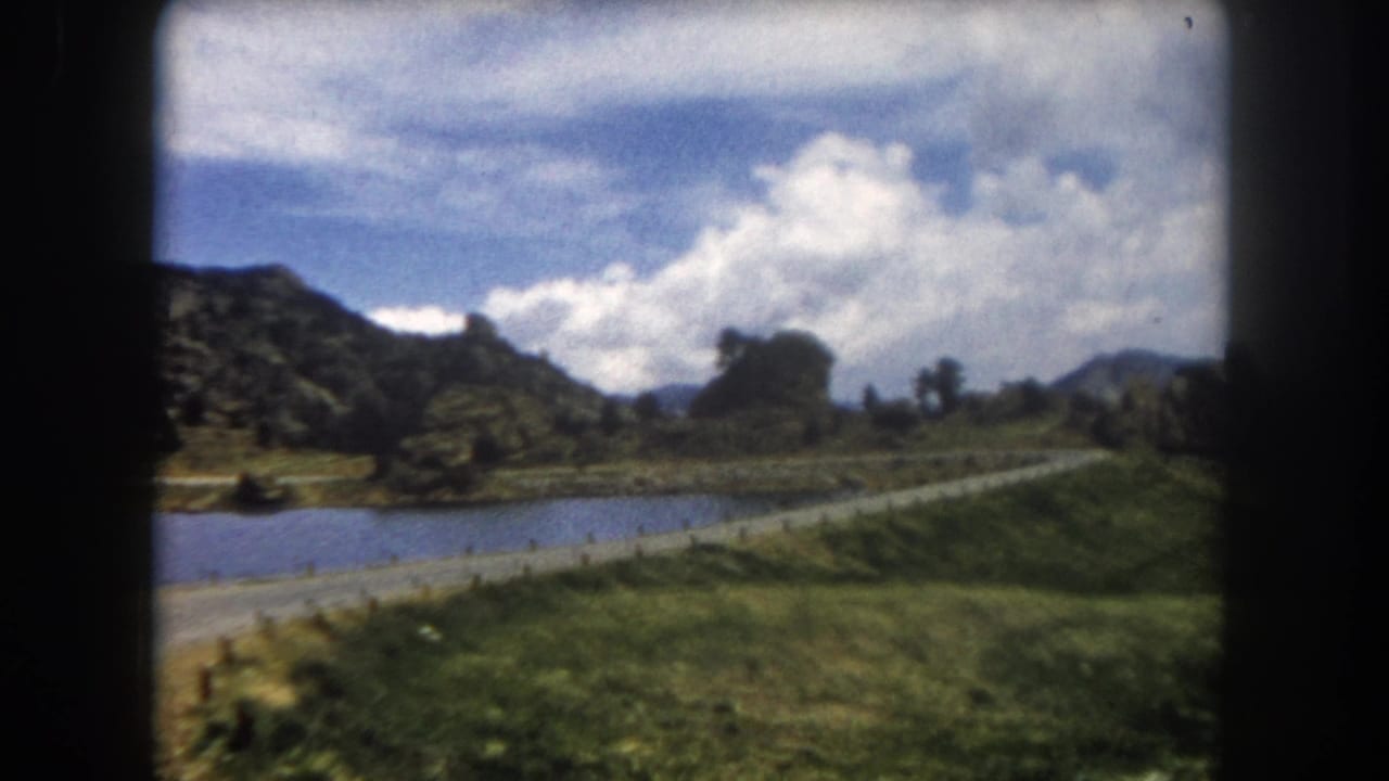 8mm film frame showing a mountain road alongside a pond with rocky peaks and cumulus clouds above a green valley