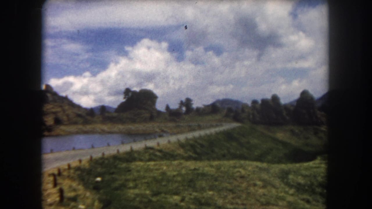 8mm film frame of a mountain valley with road, pond, wooden fence posts, rocky formations, and blue sky with white clouds