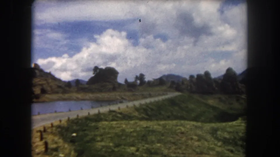 8mm home movie frame, Summit County, Colorado, circa 1962: road with wooden fence posts, a pond to the left, rugged rocky peaks, and a blue sky with white cumulus clouds