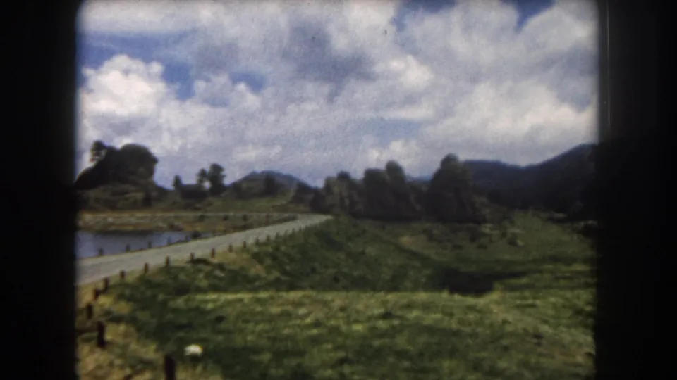 8mm home movie frame, Summit County, Colorado, circa 1962: camera pans across a green alpine meadow with a road, wooden rail fence, and rocky outcrops at mid-distance