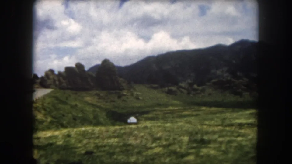 8mm home movie frame, Summit County, Colorado, circa 1962: broad green valley floor with a mountain road, rocky ridgeline, and a small white object of uncertain nature in the meadow foreground