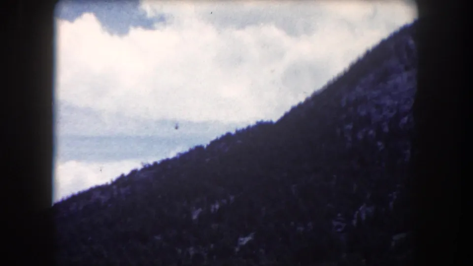 8mm film still circa 1962 showing a dark coniferous ridgeline in Summit County, Colorado, angled across the frame against a bright, cloud-streaked sky.
