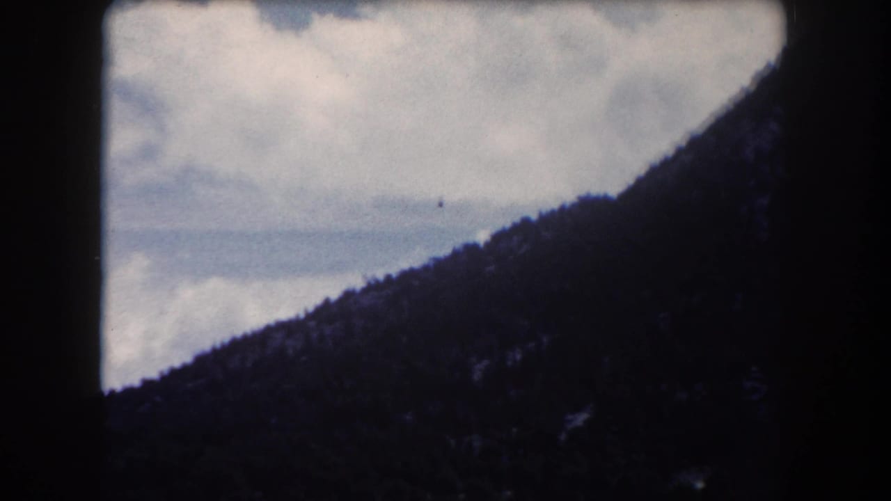 Dark forested mountain slope and ridgeline silhouetted against a cloudy sky, 8mm film with pronounced color shift.