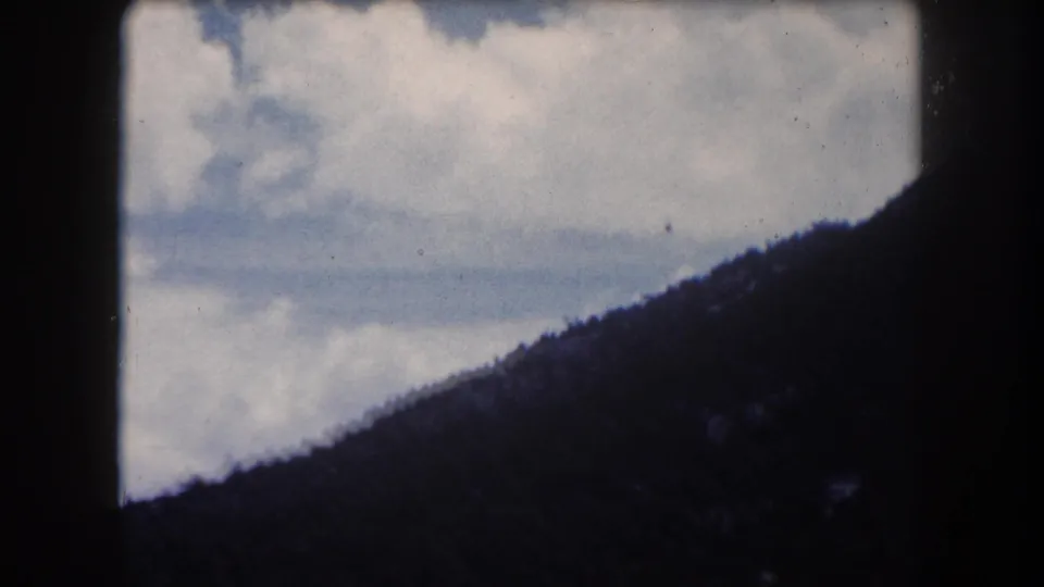 8mm home movie still circa 1962, camera panned upward to reveal large cumulus clouds above a conifer ridgeline, Summit County, Colorado.