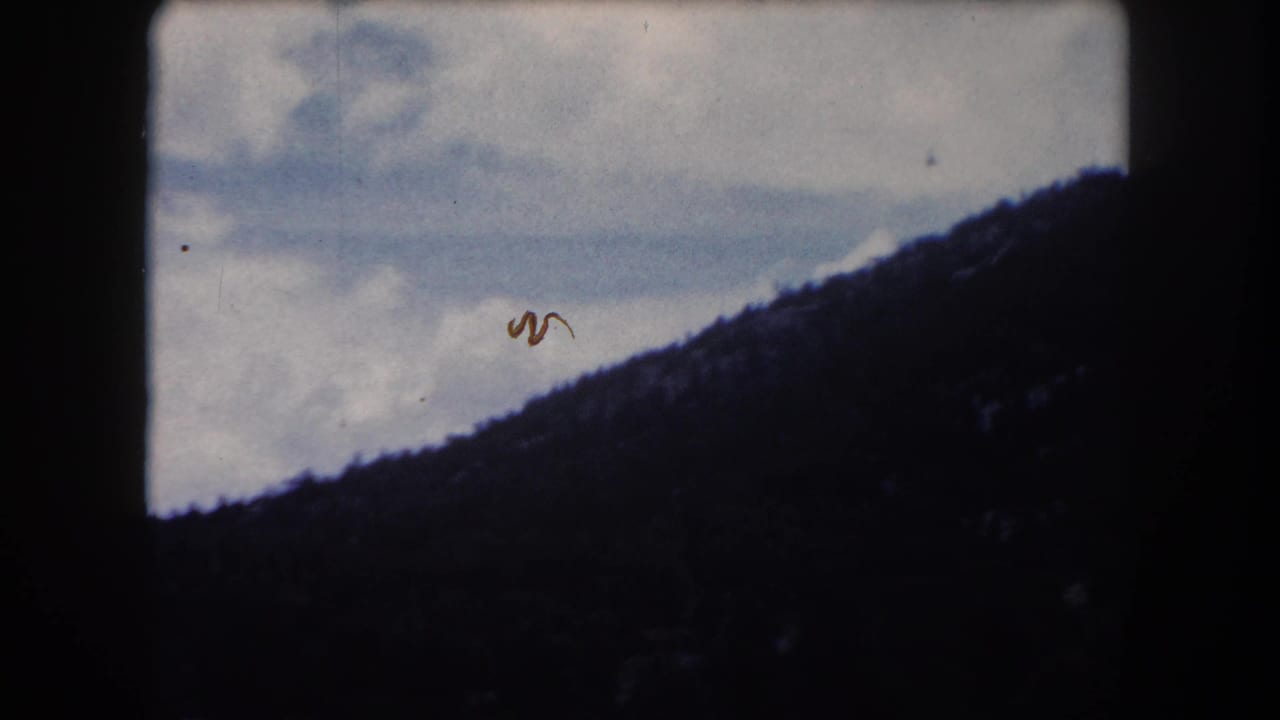 8mm home-movie frame circa 1962, Summit County Colorado: forested ridge under scattered cumulus clouds with a hair or fiber artifact in the sky