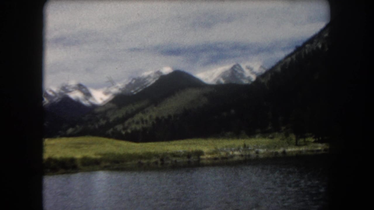 8mm film frame showing a mountain lake in the foreground, green alpine meadow, and snow-capped peaks, Summit County, Colorado, circa 1962.