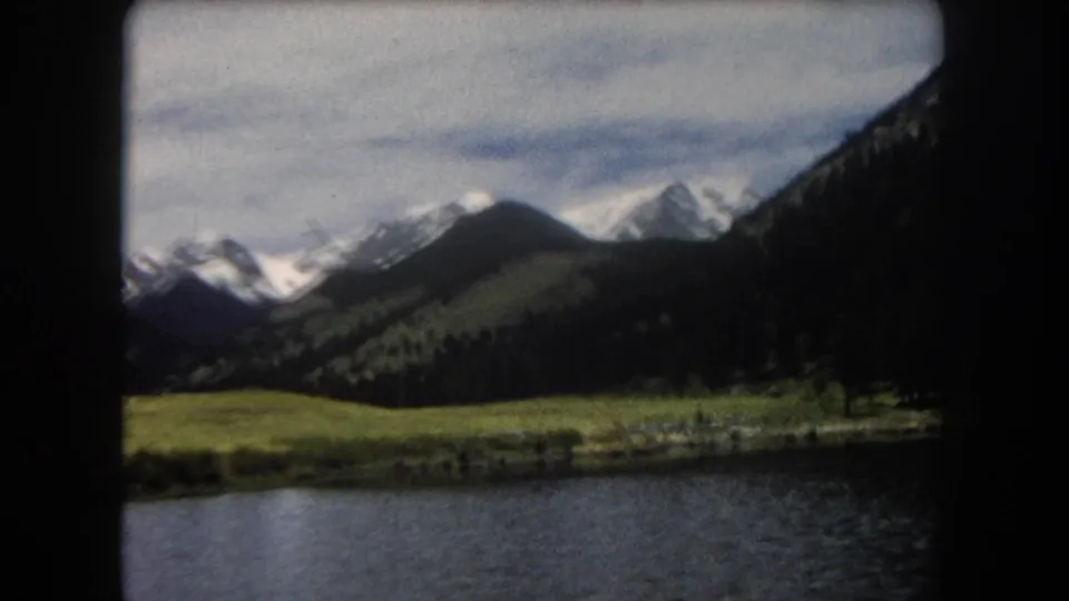 8mm film frame showing a mountain lake in the foreground, green alpine meadow, and snow-capped peaks, Summit County, Colorado, circa 1962.