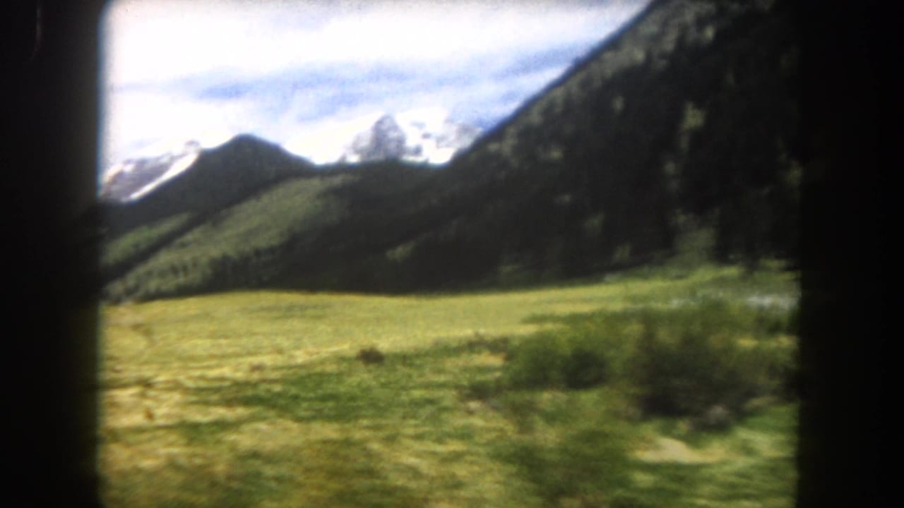 8mm film frame showing a green alpine meadow with forested hills and snow-capped mountain peaks under a partly cloudy sky, shot from a moving vehicle.