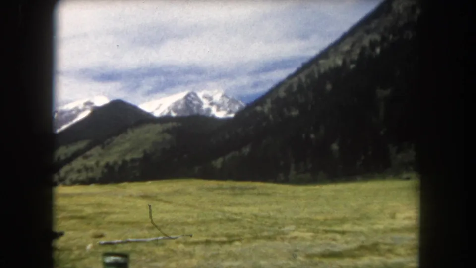 8mm home-movie frame, circa 1962, Summit County, Colorado: snow-capped Rocky Mountain peaks and forested slopes above open meadow; a small unidentified object is visible at lower left.