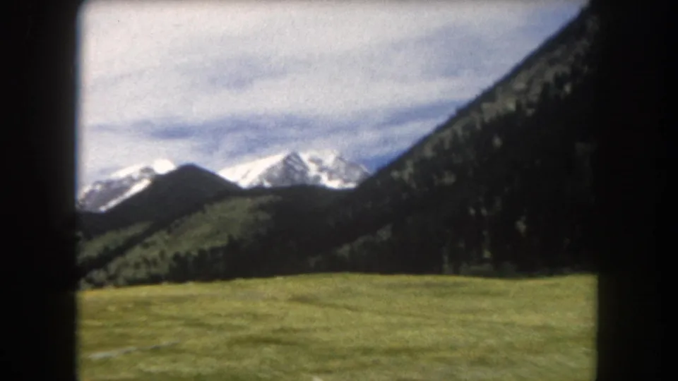 8mm home-movie frame, circa 1962, showing snow-capped peaks and conifer-forested hills above open meadow in Summit County, Colorado, with warm color cast of aged consumer film.