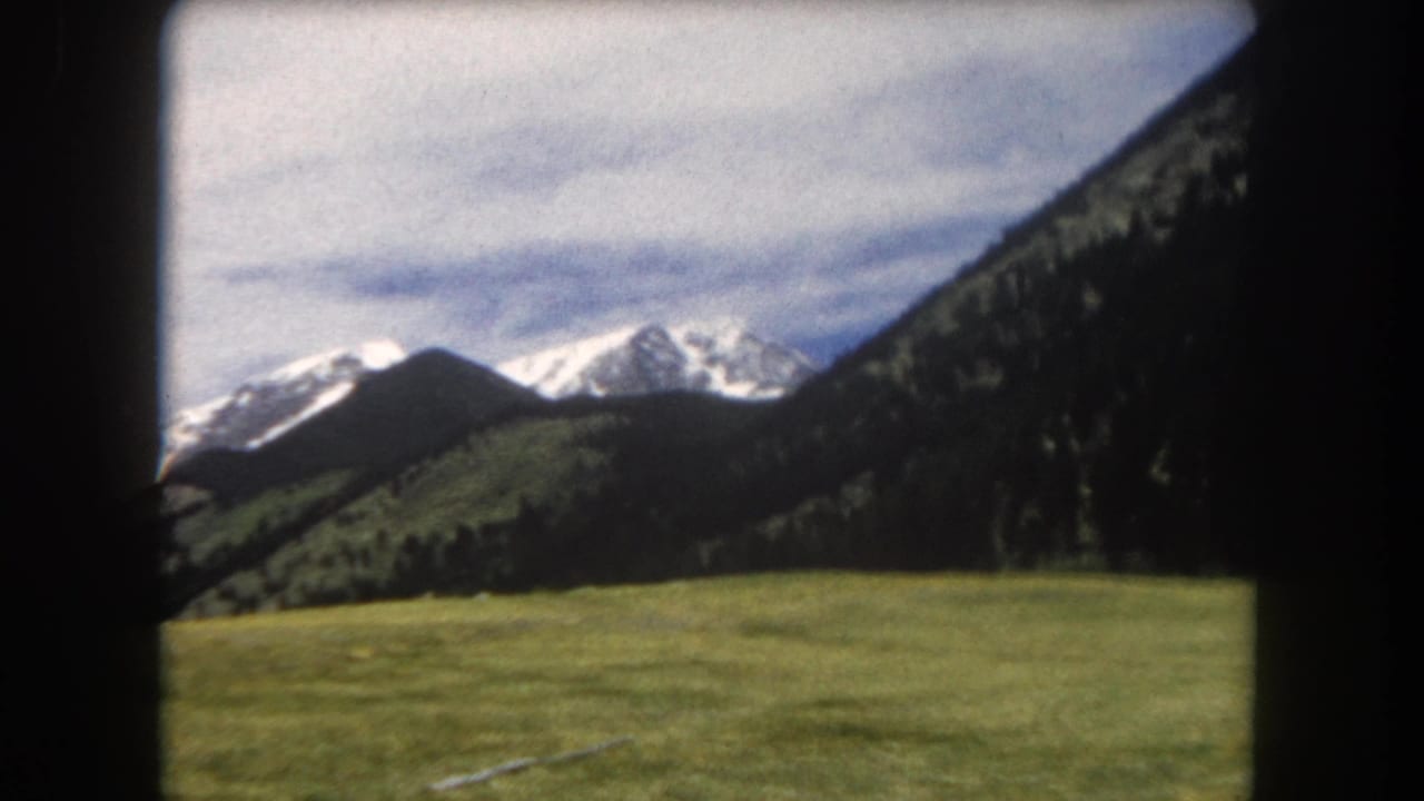 8mm color film from 1962 showing a green alpine meadow, conifer-covered mountain slopes, and snow-capped Rocky Mountain peaks in Summit County, Colorado.