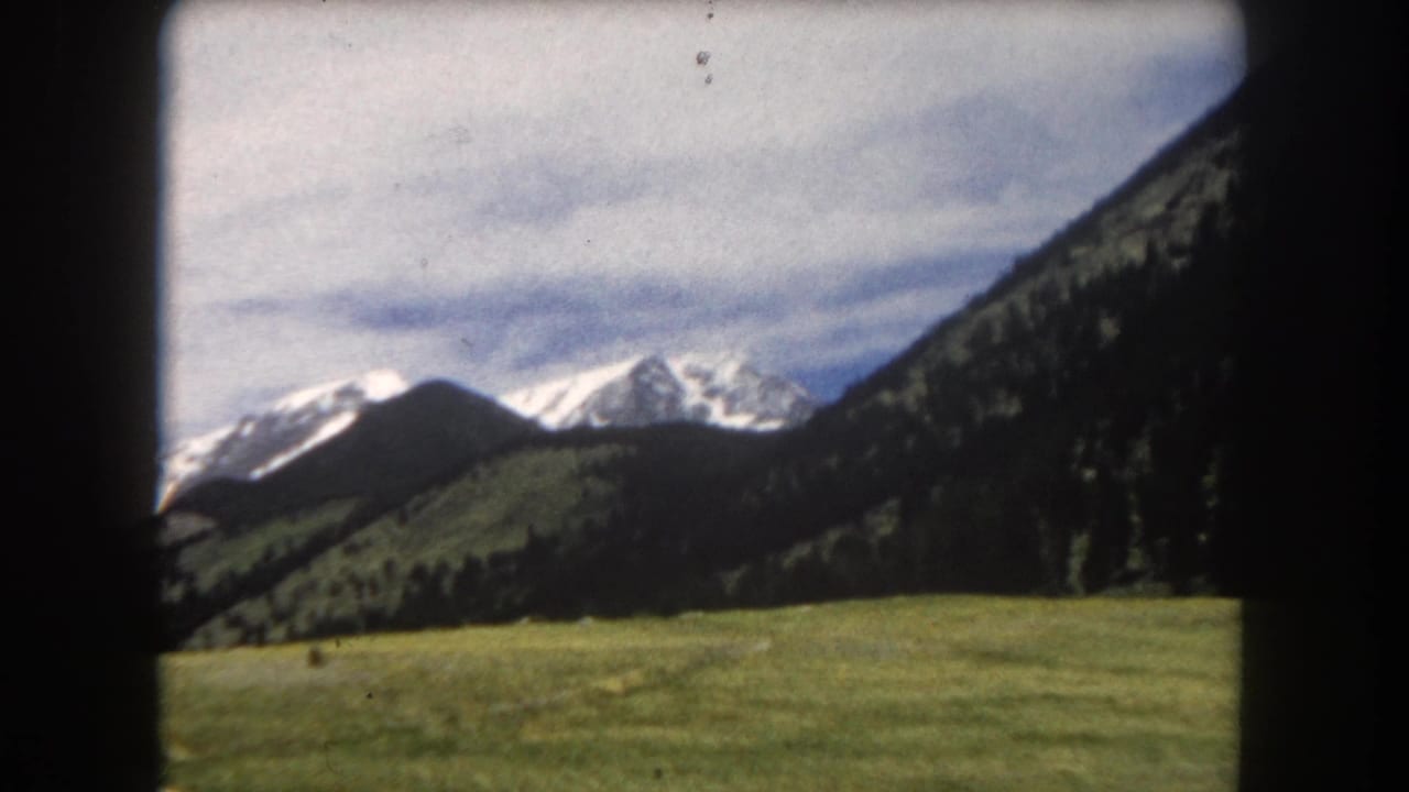 Pan continues across alpine valley; green meadow in foreground, dark forested slopes, snow-capped peaks, cloudy sky, small dark shape on the meadow.