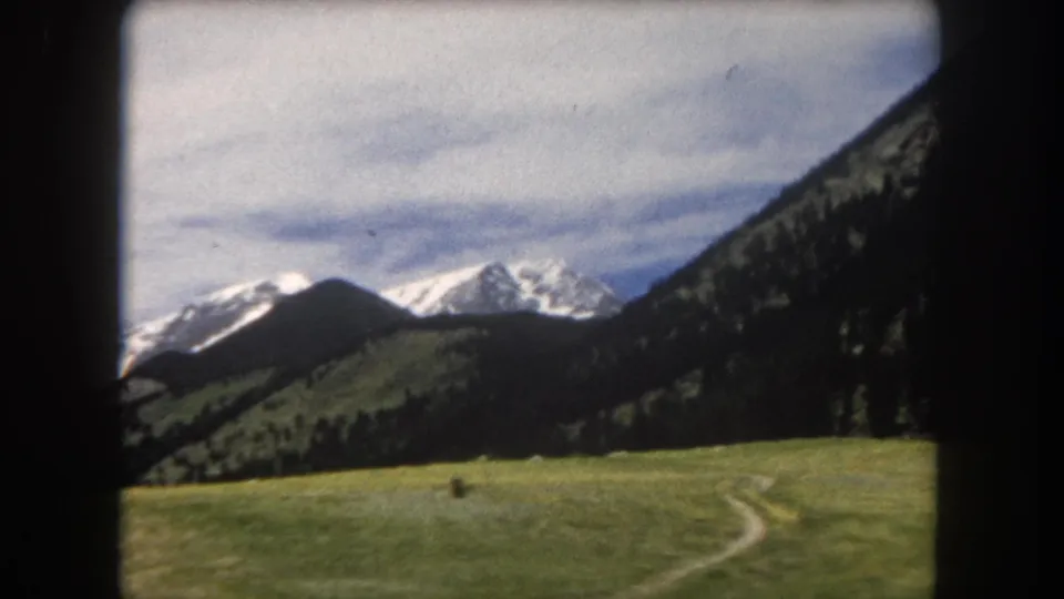 8mm color film, ca. 1962, Summit County, Colorado; a dirt path or ranch track cuts across the green alpine meadow toward conifer-covered slopes and snow-capped peaks.