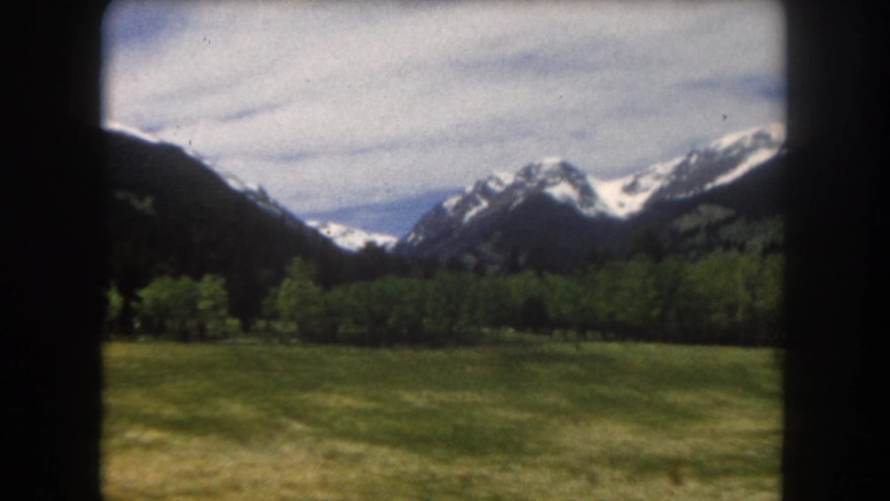 Green meadow and treeline below snow-capped peaks, shot on 8mm film with visible film-gate vignetting.