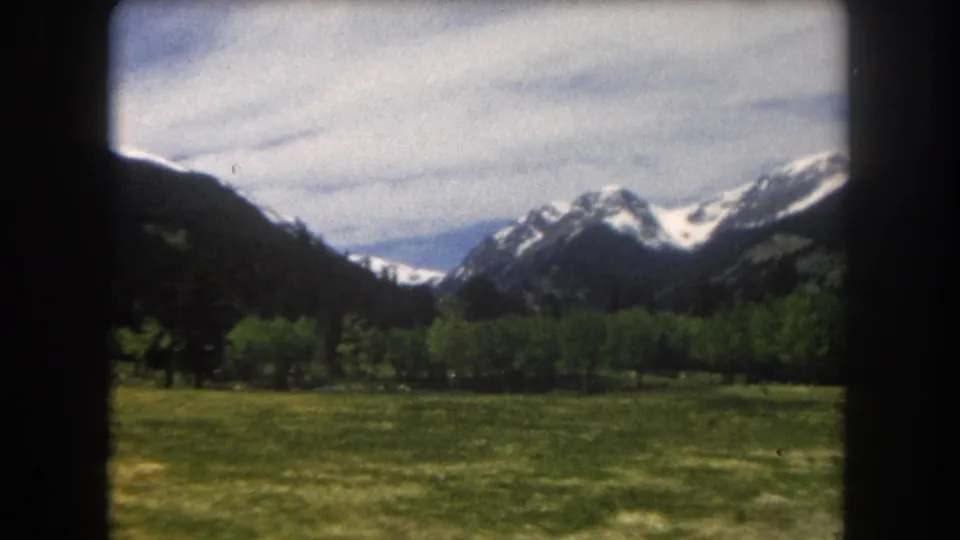 1960s 8mm home movie panning across a Colorado Rocky Mountain valley — snow-capped peaks, treeline, and green meadow, Summit County area.