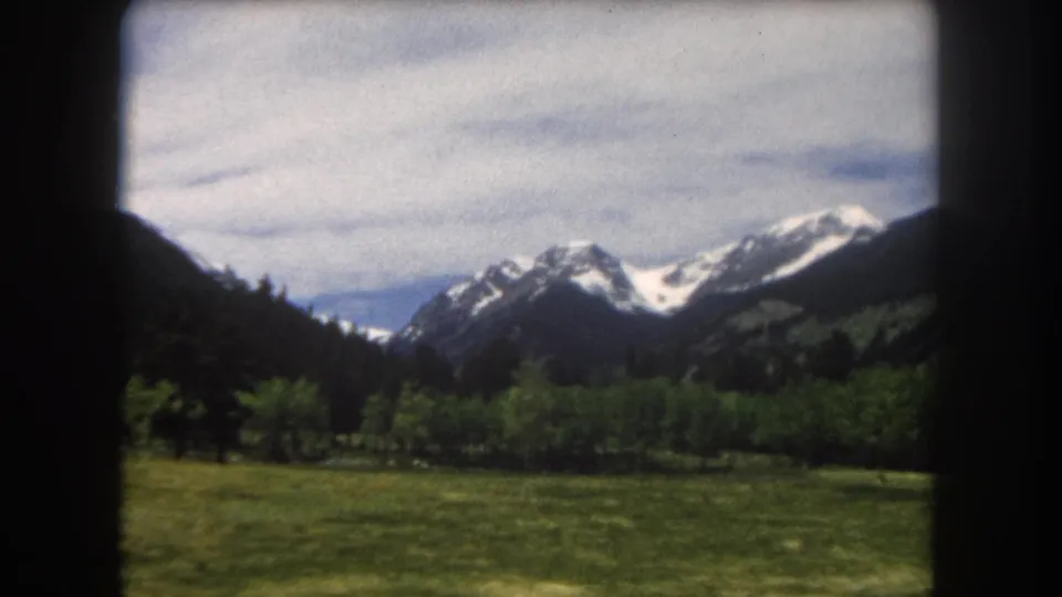 Mid-clip 8mm frame showing a Colorado mountain valley — green grass foreground, forested slopes, and snow-capped summits under an overcast sky.