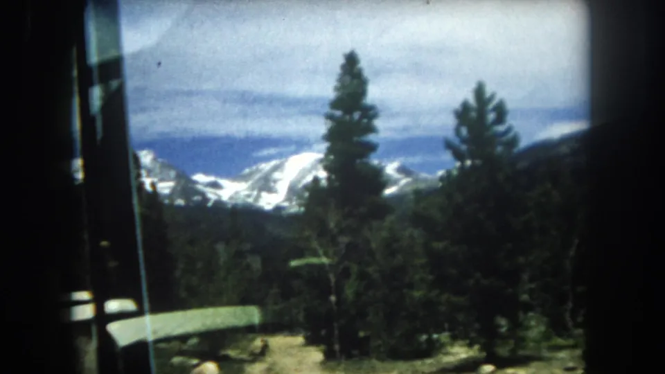 Mountain scenery shot through a vehicle window with foreground pine trees and distant snow-capped peaks, Summit County, Colorado, 8mm home movie, 1962.