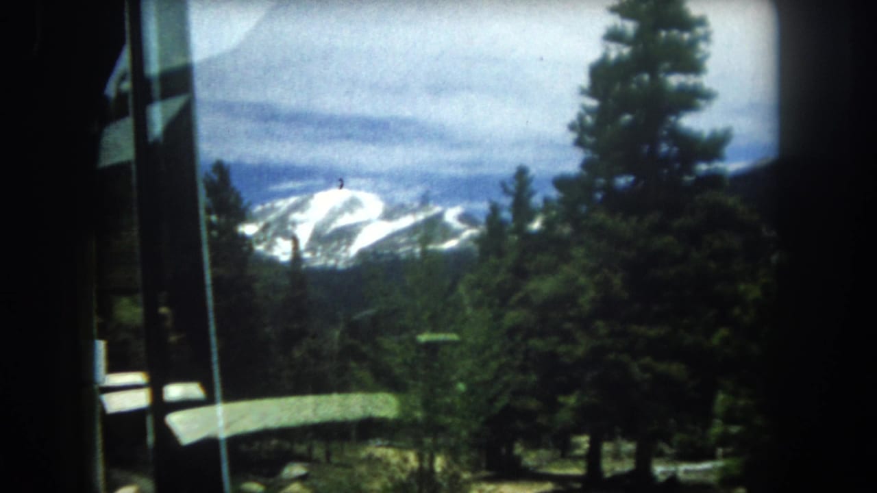 Snow-capped mountain range visible through a window, framed by pine trees and a partly cloudy sky.