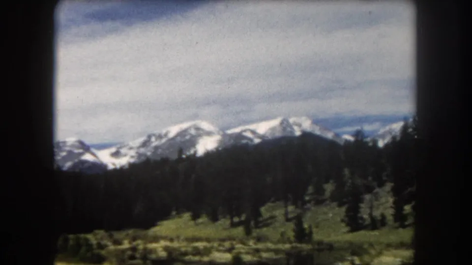8mm home movie panorama, circa 1962: snow-covered mountain range above conifer forest and alpine meadow, likely Summit County, Colorado.