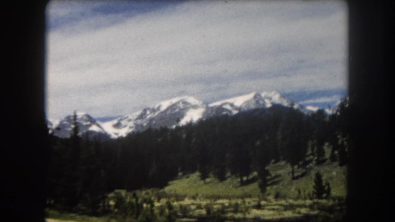8mm home-movie frame showing multiple snow-capped Rocky Mountain peaks above a conifer forest and green alpine meadow, likely Summit County, Colorado, circa 1962.