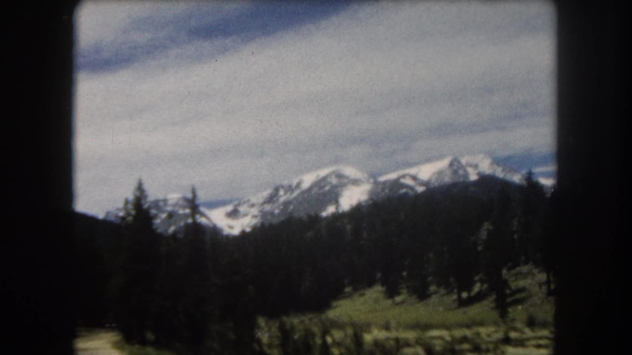 8mm home-movie frame from a moving vehicle showing a snow-covered Rocky Mountain ridgeline above dense pine forest and a green valley, Summit County, Colorado area, circa 1962.