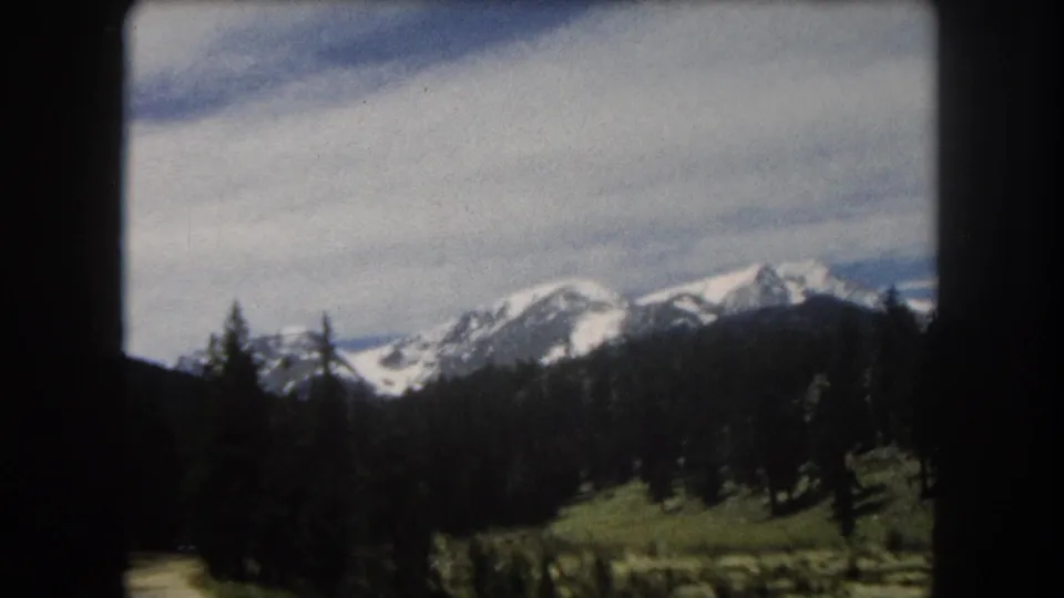 8mm home-movie frame from a moving vehicle showing a snow-covered Rocky Mountain ridgeline above dense pine forest and a green valley, Summit County, Colorado area, circa 1962.