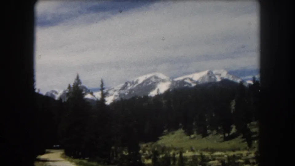8mm home-movie frame showing snow-capped alpine peaks, conifer trees, and a green meadow as the camera pans during a road trip, Summit County, Colorado area, circa 1962.