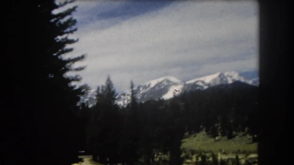 8mm home-movie frame with a tall conifer tree dominating the foreground as the vehicle moves, snow-capped mountain peaks visible beyond, Summit County, Colorado area, circa 1962.