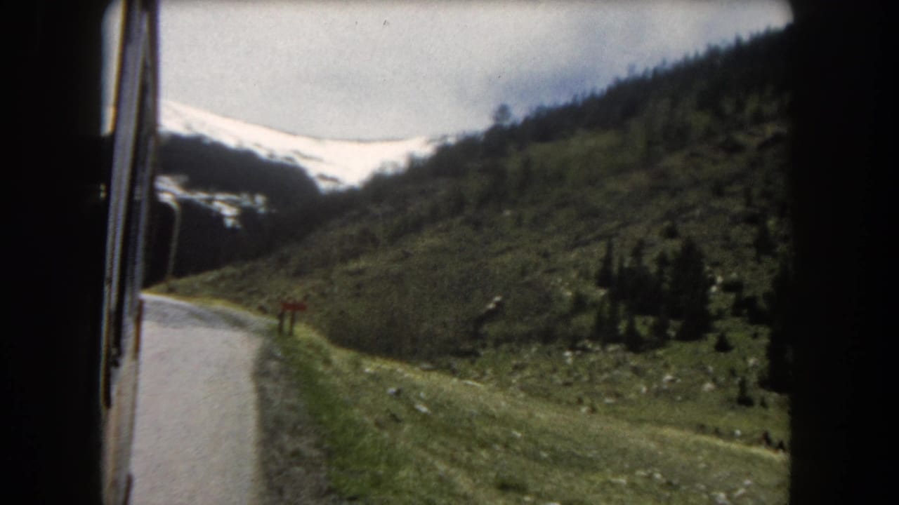 8mm home-movie frame c.1962, Summit County, Colorado: moving-vehicle POV showing a mountain road, green slopes, scattered pines, and snow on distant peaks.