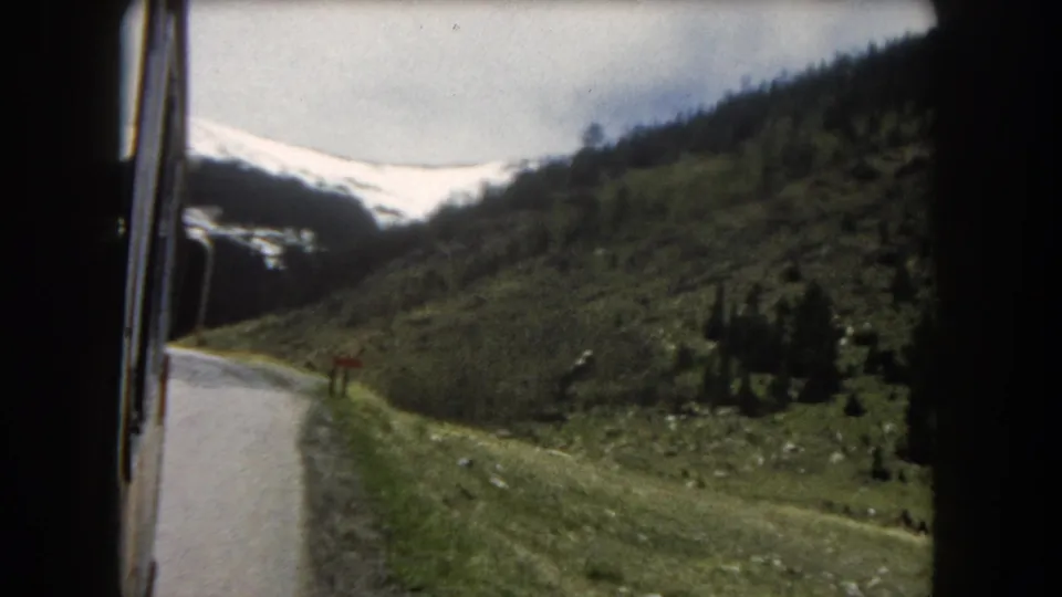 8mm home-movie frame c.1962, Summit County, Colorado: moving-vehicle POV showing a mountain road, green slopes, scattered pines, and snow on distant peaks.