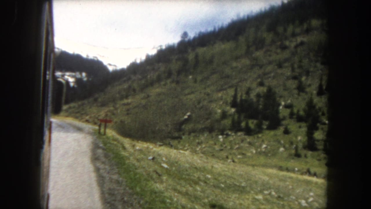 Moving-vehicle window view of a winding mountain road with conifer-covered green slopes and snow patches visible on the upper ridgeline.