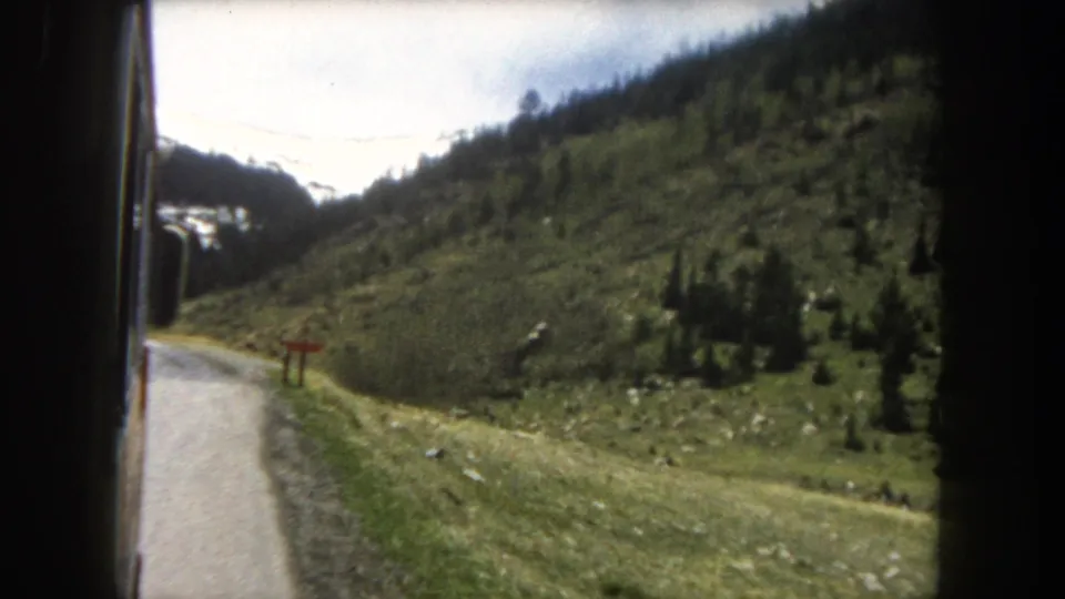 8mm home-movie frame c.1962, Summit County, Colorado: vehicle POV on a mountain road showing a winding surface, green hillsides, conifers, ridgeline snow, and red roadside marker.