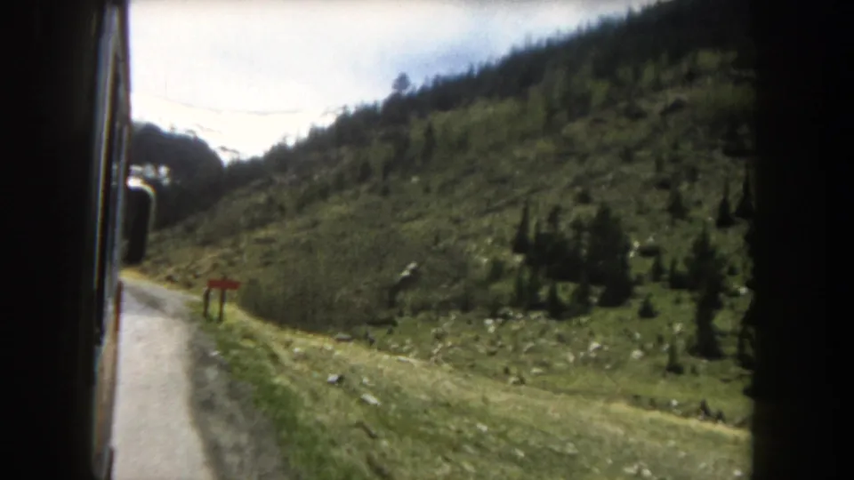 8mm home-movie frame c.1962, Summit County, Colorado: moving vehicle shows pine-covered mountain slope, road ahead, red markers along verge, and utility lines overhead.