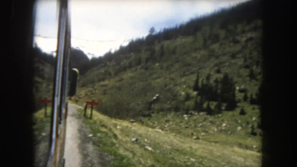 8mm home-movie frame c.1962, Summit County, Colorado: vehicle POV continuing through mountain terrain with green slopes, conifers, red roadside markers, and utility lines.