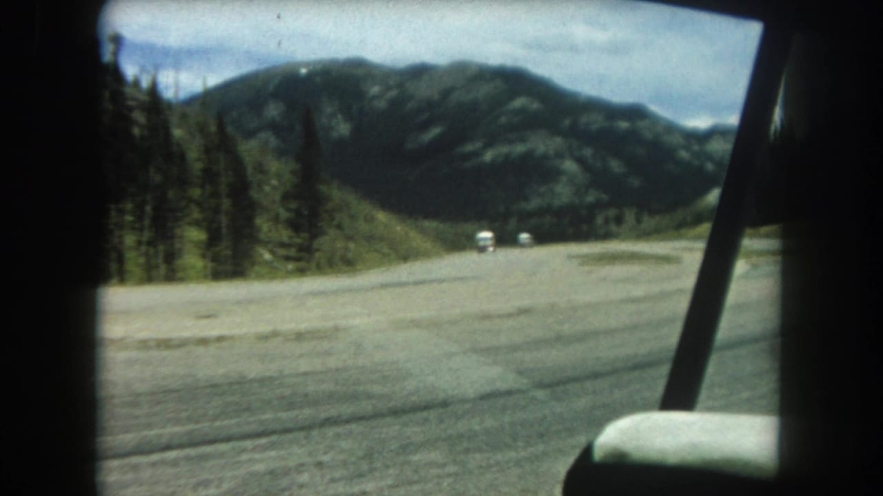 View through car window of a wide mountain road with two white vehicles ahead and conifer-covered slopes beyond.