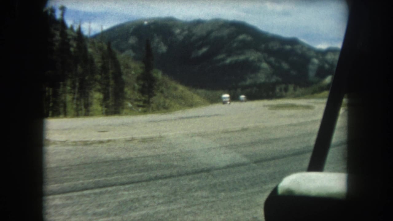 In-car POV of a wide road through conifer forest with mountain ridges and two small white vehicles ahead.